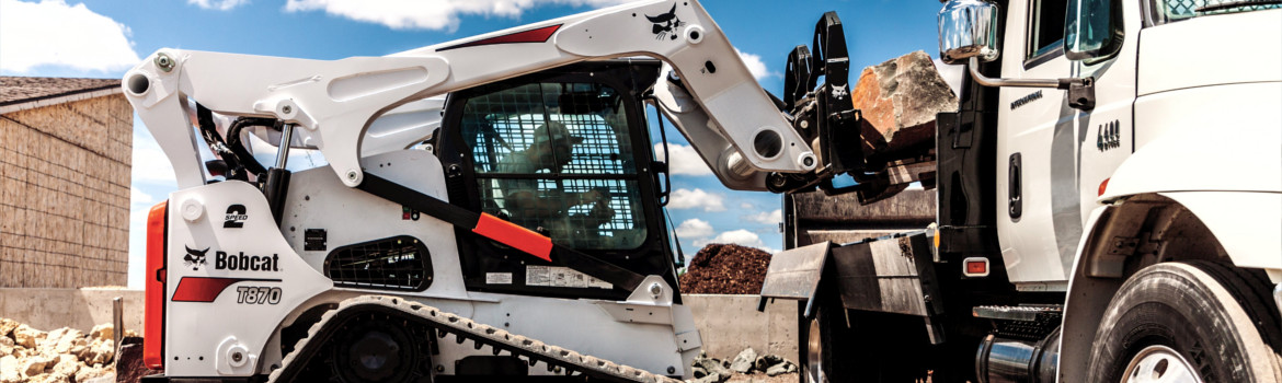 Man operating Bobcat® compact track loader by dumping large boulder into truck bed on work site
